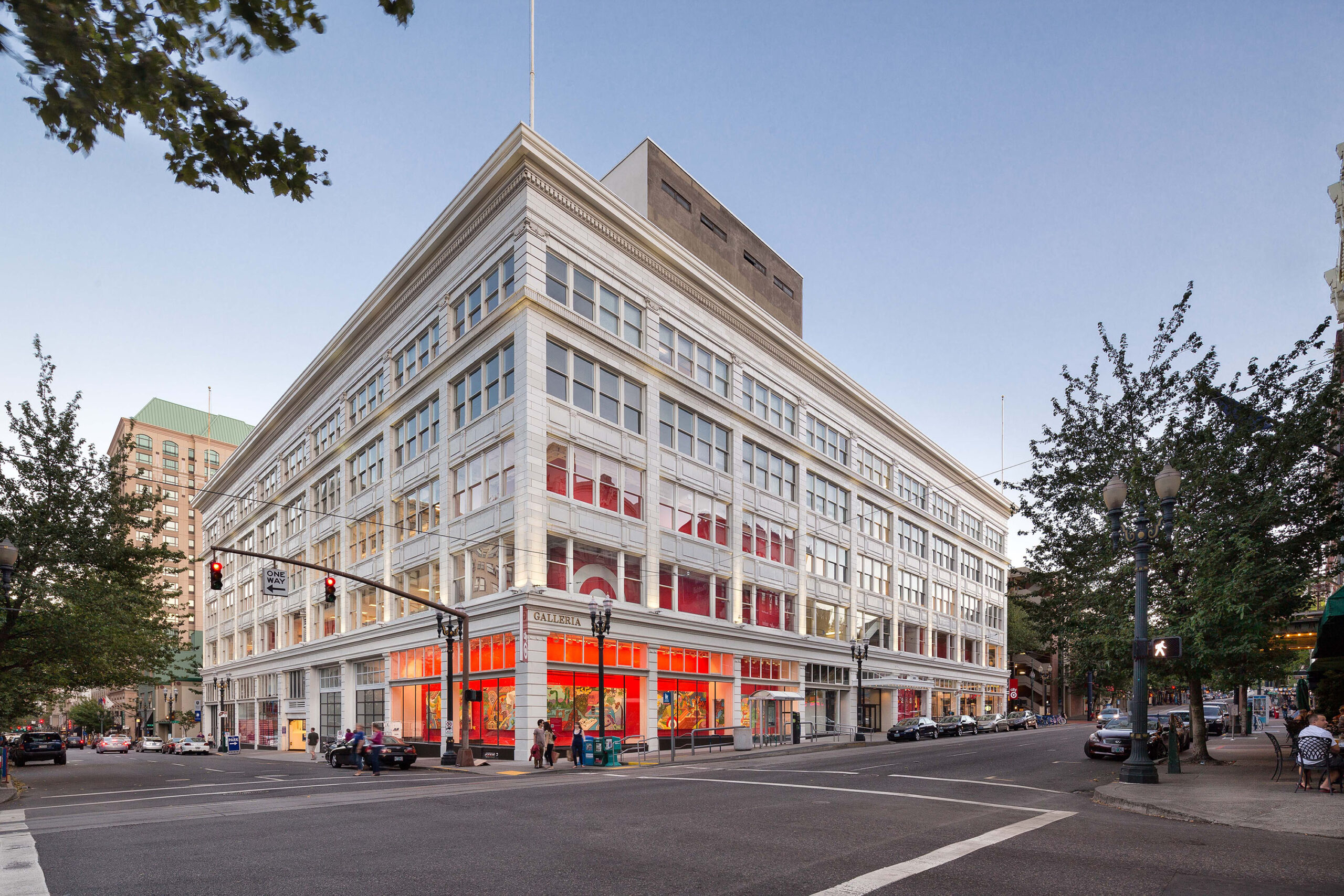 The Galleria Building in Portland, Oregon, a historic multi-story structure with a white façade and large windows. The corner of the building features a Target store with red signage and bright interior lighting visible from the street. The urban setting includes traffic lights, pedestrians, and surrounding buildings under a clear evening sky